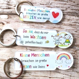 Könnte beinhalten: Drei rechteckige Acryl-Schlüsselanhänger mit Metallringen. Der oberste trägt die Aufschrift "Besondere Menschen brauchen so große HERZEN wie deins!" mit einem roten Herz. Der mittlere sagt "Danke, dass du mir beim Wachsen geholfen hast!" mit zwei Kindern und einem Regenbogen. Der untere zeigt "Kleine Menschen brauchen so große Herzen wie Deins" mit einem Regenbogen.