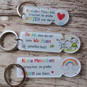 May include: Three rectangular acrylic keychains with metal rings. The top one reads "Besondere Menschen brauchen so große HERZEN wie deins!" with a red heart. The middle one says "Danke, dass du mir beim Wachsen geholfen hast!" with two children. The bottom one says "Kleine Menschen brauchen so große Herzen wie Deins" with a rainbow.