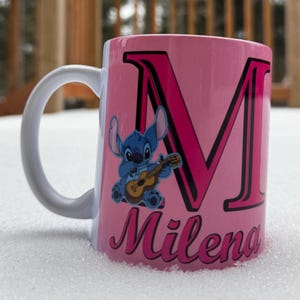 Op de afbeelding: Een roze keramische mok met een witte handgreep. De mok heeft een blauw cartoonfiguur dat gitaar speelt, een grote roze letter "M" met een zwarte omtrek en de naam "Milena" in roze cursief.