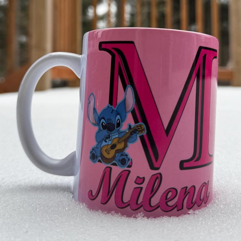 Op de afbeelding: Een roze keramische mok met een witte handgreep. De mok heeft een blauw cartoonfiguur dat gitaar speelt, een grote roze letter "M" met een zwarte omtrek en de naam "Milena" in roze cursief.