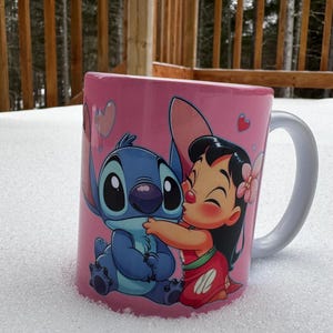 Op de afbeelding: Een roze keramische mok met een cartoon afbeelding van Stitch en Lilo uit de Disney-film Lilo & Stitch. Stitch is blauw en heeft grote ogen. Lilo draagt een rood-witte jurk en heeft een bloem in haar haar. De mok heeft een wit handvat.