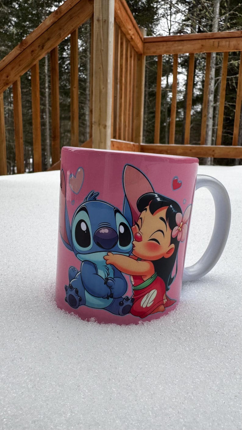 Op de afbeelding: Een roze keramische mok met een cartoon afbeelding van Stitch en Lilo uit de Disney-film Lilo & Stitch. Stitch is blauw en heeft grote ogen. Lilo draagt een rood-witte jurk en heeft een bloem in haar haar. De mok heeft een wit handvat.
