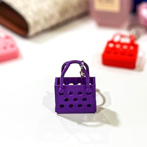 A purple 3D printed handbag keychain with a perforated design, displayed on a white surface. Background includes other handbag keychains in red and pink, along with accessories like perfume and a wallet.