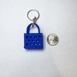 A blue tote bag keychain made out of pla plus plastic laying flat next to a quarter to compare sizes. It is fairly taller than a quarter but not much wider.