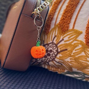 Pumpkin keychain clipped to tan handbag with patterned fall blanket – spooky cute Halloween charm.