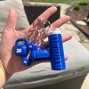 A set of blue keychain charms with a gym theme. The charms include a dumbbell, a kettlebell, a gym bag, and a water bottle. The kettlebell has the number 20 on it, the dumbbell has the number 50 on it, and the gym bag has the word 'GAINS' on it. The water bottle has the word 'HYDRATE' on it.