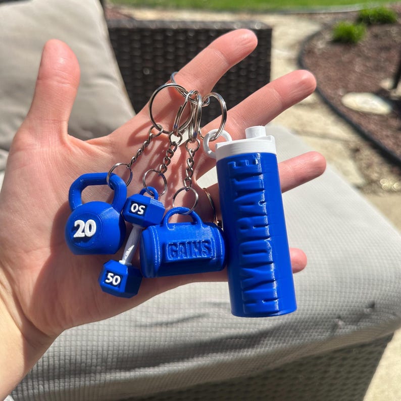A set of blue keychain charms with a gym theme. The charms include a dumbbell, a kettlebell, a gym bag, and a water bottle. The kettlebell has the number 20 on it, the dumbbell has the number 50 on it, and the gym bag has the word 'GAINS' on it. The water bottle has the word 'HYDRATE' on it.