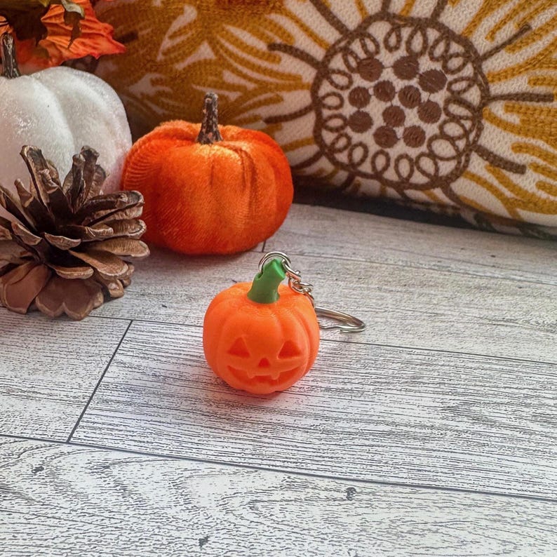 Pumpkin Halloween keychain displayed with small pumpkins and pinecones on rustic wood background.