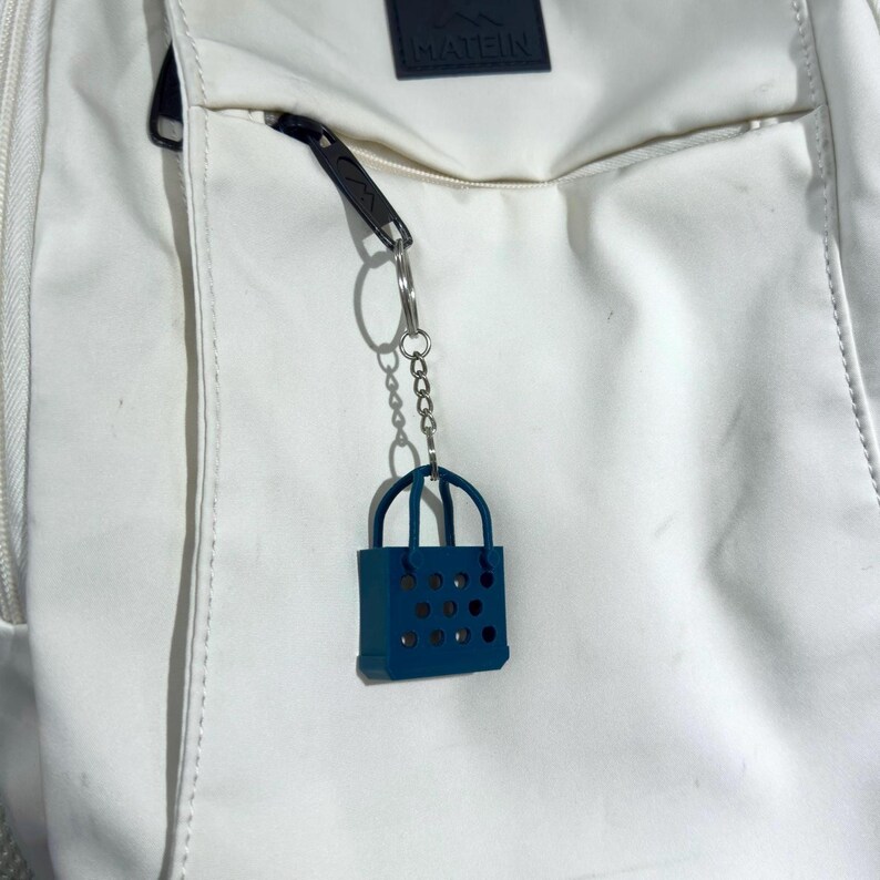A dark green 3D printed handbag keychain with a perforated design, attached to the zipper of a white backpack. The bag adds a stylish, playful accessory to the clean, minimal background.