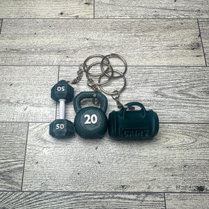A green dumbbell keychain with a 50 on it, a green kettlebell keychain with a 20 on it, and a green gym bag keychain with the word 'GAINS' on it. All three keychains are attached to a silver key ring.