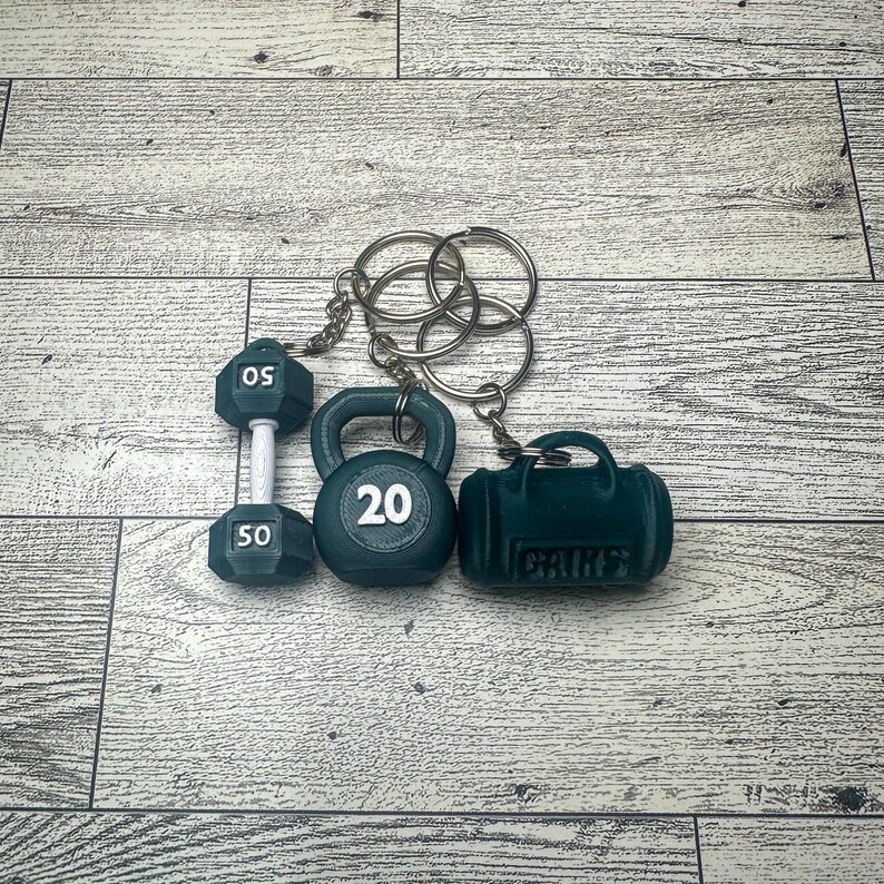 A green dumbbell keychain with a 50 on it, a green kettlebell keychain with a 20 on it, and a green gym bag keychain with the word 'GAINS' on it. All three keychains are attached to a silver key ring.