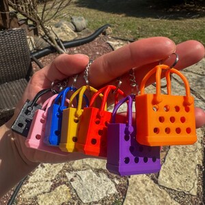 Seven of the mini tote bag keychains in various colors including orange, black, pink, blue, yellow, red, and purple. The keychains are made of PLA Plus plastic material. The bag handles are connected to be able to attach any other ring sizes not provided.