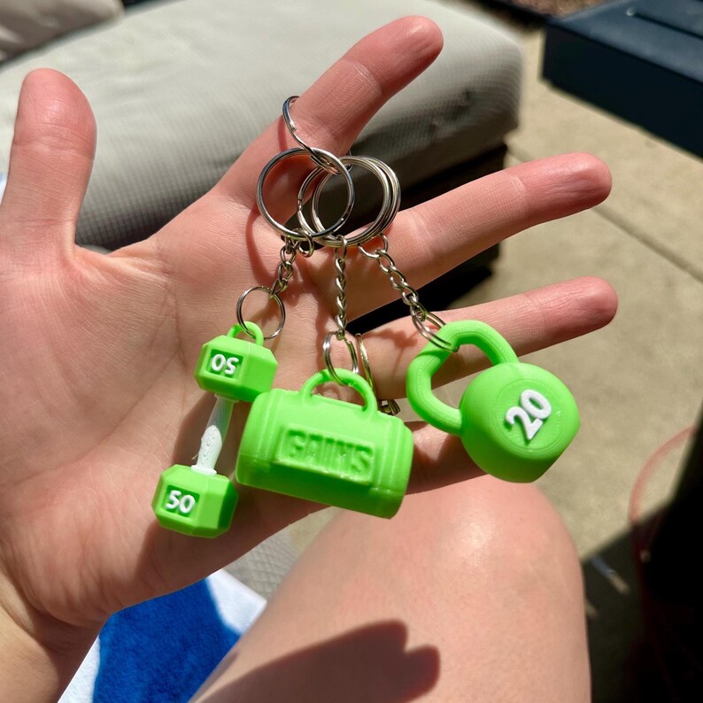 A set of four green keychain charms: a dumbbell with '50' printed on it, a gym bag with 'GAINS' printed on it, a kettlebell with '20' printed on it, and another dumbbell with '50' printed on it.
