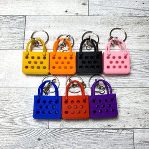 Seven of the different colors of the mini tote bag keychain. Clean professional image of the keychains on a white monochrome wood surface.