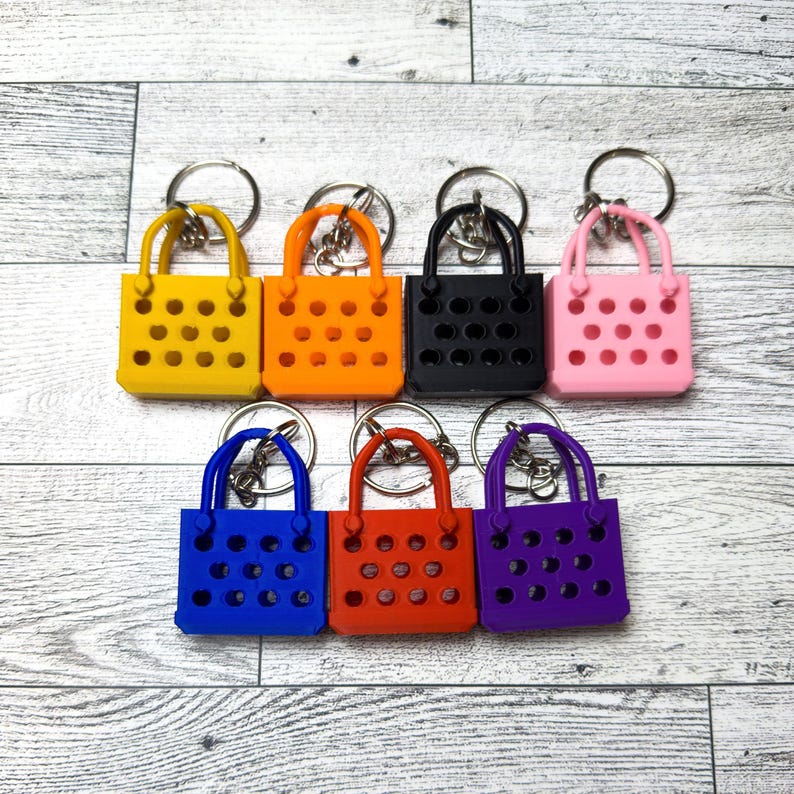 Seven of the different colors of the mini tote bag keychain. Clean professional image of the keychains on a white monochrome wood surface.