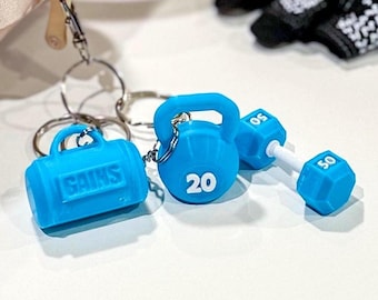 May include: A set of blue fitness-themed keychains. The keychains include a miniature gym bag with the word "GAINS", a kettlebell with the number "20", and a dumbbell with "50" on each end. The keychains are attached to silver keyrings.