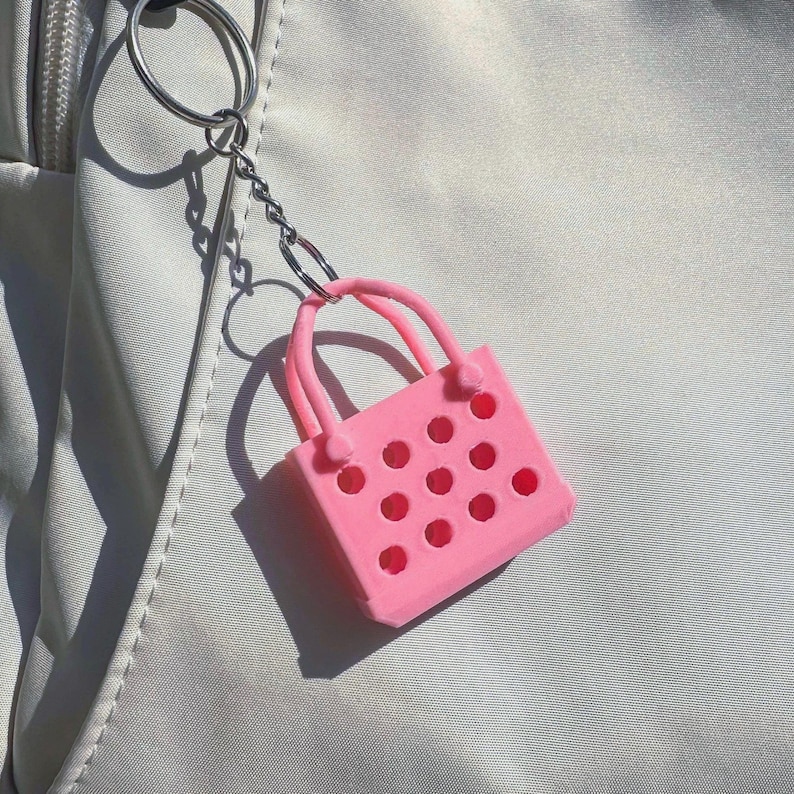 A pink plastic keychain shaped like a small shopping bag with a silver chain. The bag has a handle and a pattern of small circles on the front.