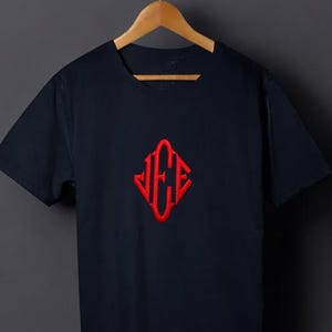 Könnte beinhalten: Marineblaues T-Shirt mit rotem, gesticktem Diamant-Design und den Buchstaben "JCE". Das T-Shirt hängt an einem Holzbügel vor einem dunkelgrauen Hintergrund.
