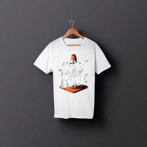 May include: White t-shirt with the text "Hoist the Cone" in a stylized font, featuring an orange traffic cone graphic. The shirt is displayed on a wooden hanger against a dark grey background. The design is centered on the shirt.