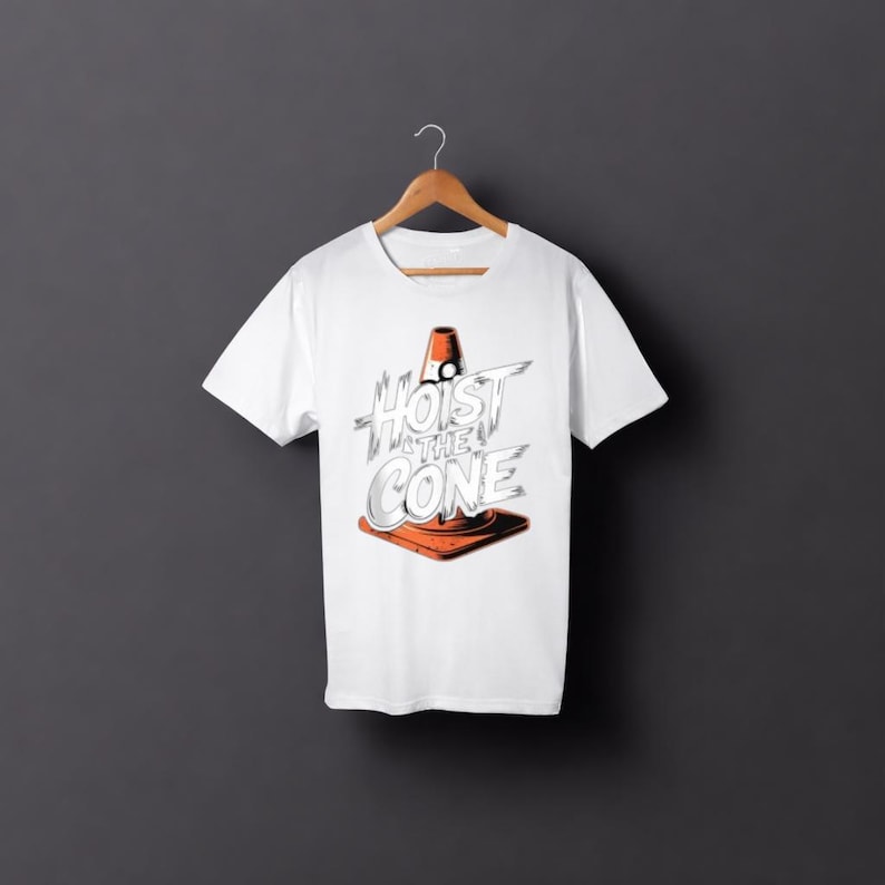 May include: White t-shirt with the text "Hoist the Cone" in a stylized font, featuring an orange traffic cone graphic. The shirt is displayed on a wooden hanger against a dark grey background. The design is centered on the shirt.