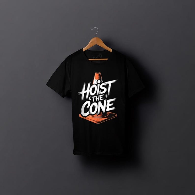 May include: Black t-shirt featuring the text "Hoist the Cone" in white, with an orange traffic cone graphic. The shirt is displayed on a wooden hanger against a dark grey backdrop.
