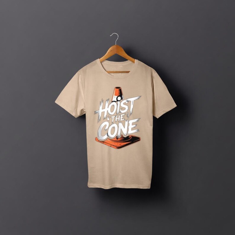 May include: A beige t-shirt displaying the phrase "Hoist the Cone" in white and grey, accompanied by an orange traffic cone illustration. The tee is suspended on a wooden hanger against a grey backdrop.