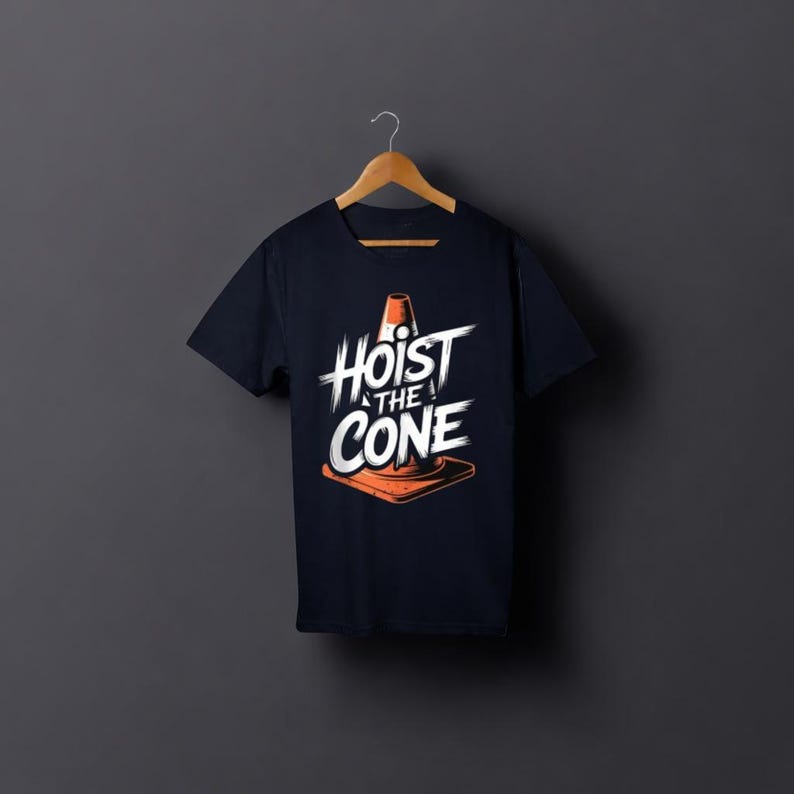 May include: Dark navy blue t-shirt featuring the text "Hoist the Cone" in white, with an orange traffic cone graphic. The tee shirt is displayed on a wooden hanger against a dark grey backdrop.