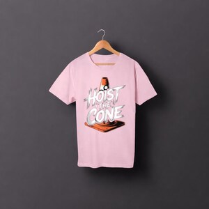 May include: A pale pink t-shirt with the words "Hoist the Cone" in a graphic design featuring a traffic cone. The design is in white, orange, and black. The shirt is hanging on a wooden hanger.