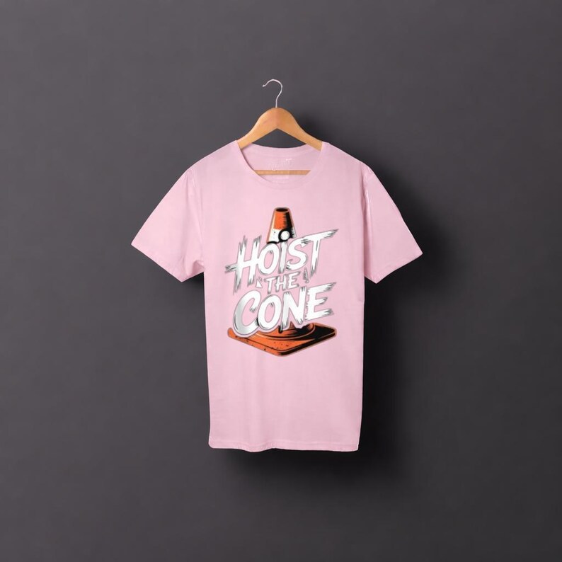 May include: A pale pink t-shirt with the words "Hoist the Cone" in a graphic design featuring a traffic cone. The design is in white, orange, and black. The shirt is hanging on a wooden hanger.