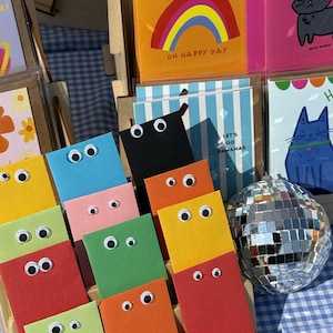 May include: A display of colorful greeting cards and stationery. The cards feature various designs, including a rainbow with the text "OH HAPPY DAY", cartoon cats, and striped patterns. Some cards have googly eyes attached. A small disco ball is also present.