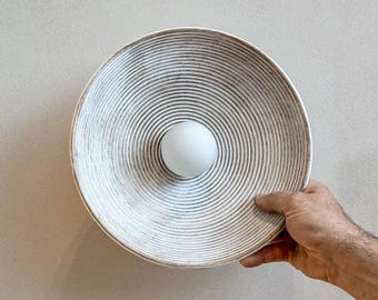 May include: A circular wall sconce featuring a wide, shallow, bowl-shaped shade with a textured, concentric ribbed pattern in off-white and grey tones, centered with a smooth, opaque white spherical light bulb.