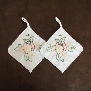 May include: Two white, square pot holders with embroidered strawberry and daisy designs. Each pot holder has a loop for hanging. The strawberries are red with yellow seeds, and the daisies are white with yellow centers.