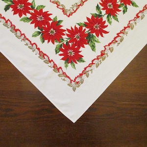 May include: A white tablecloth with a Christmas poinsettia floral design. The corners of the tablecloth are visible, showcasing the red poinsettia flowers with green leaves and a decorative border in red, gold, and white.