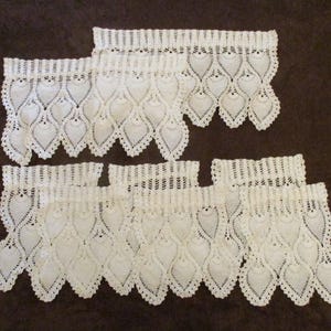 May include: A set of off-white crochet valances featuring a repeating pineapple-inspired pattern with a scalloped bottom edge. Each piece has a straight top header with a series of vertical stitches, transitioning into an intricate openwork design.