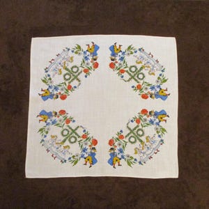 May include: White linen handkerchief with a colorful floral and figure design. The corners feature a repeating pattern of blue, yellow, and red figures, flowers, and greenery. The handkerchief is square and appears to be vintage.