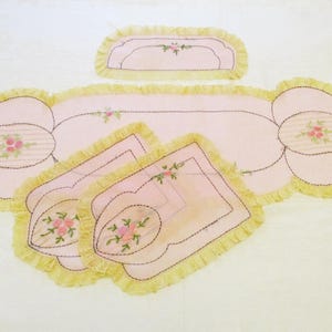 May include: A set of vintage pink and yellow embroidered table linens. The set includes a long runner, a small runner, and several placemats. Each piece features a floral design and a ruffled edge.