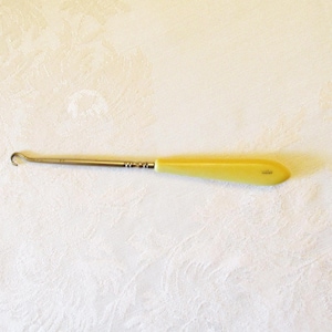 May include: A vintage crochet hook with a yellow handle and a silver metal shaft. The hook is curved at the end. The handle is a light yellow color. The hook is laying on a white, textured surface.