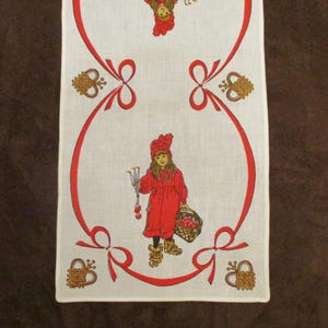May include: White rectangular table runner with a repeating pattern of a girl in a red dress, red bows, and decorative elements. The girl is holding a basket of fruit and a bundle of flowers. The runner has a red border.
