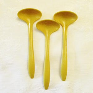 May include: Three vintage yellow serving spoons with a rounded head and long, slender handle. The spoons are arranged vertically against a white background. The spoons have a retro aesthetic.