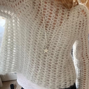 May include: A pale grey, open-knit jumper with long sleeves. The jumper has a V-neck and is worn with a silver necklace with a star pendant. The jumper is worn over a white top.
