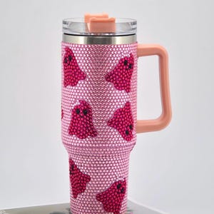 May include: A pink, bedazzled travel mug with a handle and lid. The mug is covered in small, pink rhinestones and features a pattern of red ghost-shaped designs. The handle and lid accents are a matching peach colour.