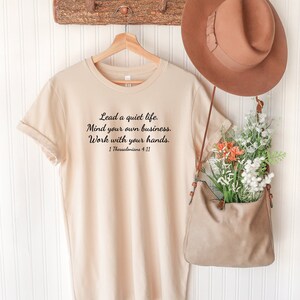 Puede incluir: Una camiseta beige con una cita en negro que dice "Lead a quiet life. Mind your own business. Work with your hands. 1 Thessalonians 4:11".