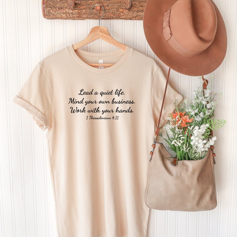 Puede incluir: Una camiseta beige con una cita en negro que dice "Lead a quiet life. Mind your own business. Work with your hands. 1 Thessalonians 4:11".