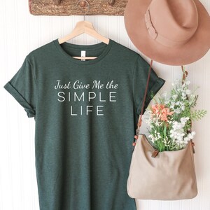 May include: A dark green t-shirt with white text that reads "Just Give Me the SIMPLE LIFE". The shirt is hanging on a wooden hanger with a brown hat and a beige bag hanging next to it.