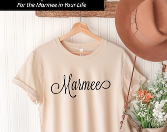 Può includere: Una t-shirt a maniche corte color beige chiaro con la parola Marmee scritta in corsivo nero sul petto. Il capo è appeso a una gruccia di legno, accompagnato da un cappello di feltro marrone e una borsa contenente fiori bianchi e arancioni.
