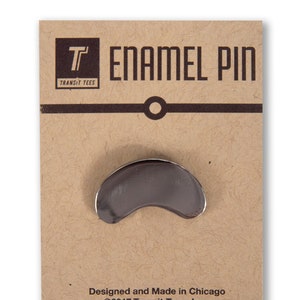 May include: A silver enamel pin shaped like a kidney bean. The pin is on a brown card with the text "T Enamel Pin Transit Tees" and "Designed and Made in Chicago ©2017 Transit Tees, Inc."