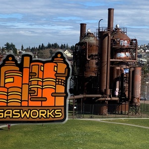 K&ouml;nnte beinhalten: Ein Aufkleber mit einer stilisierten Illustration des Gas Works Park in Seattle, Washington. Der Aufkleber hat ein braunes und orangefarbenes Farbschema mit dem Wort "GASWORKS" in einer fetten Schriftart. Der Hintergrund zeigt den Park und die Stadt.