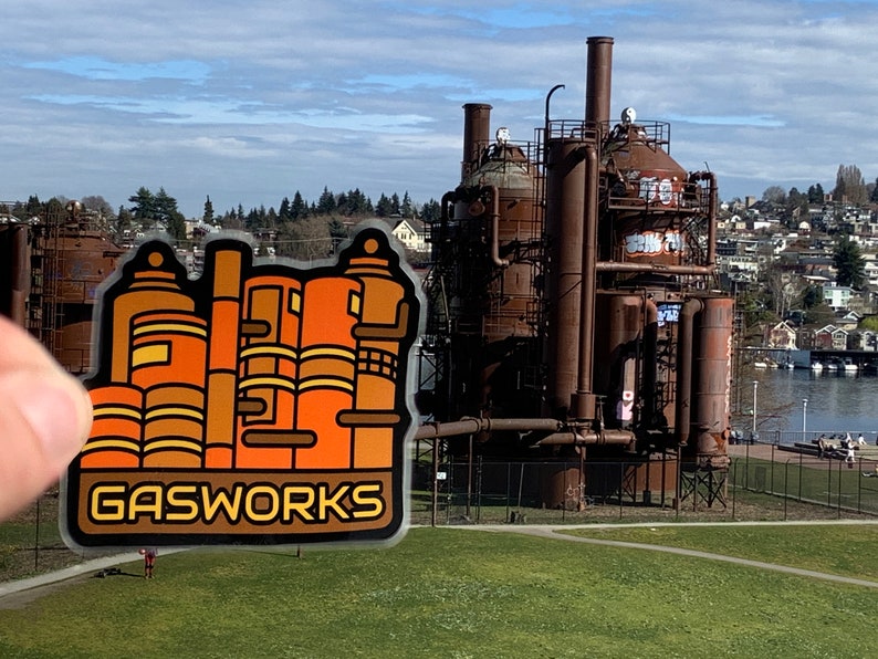 K&ouml;nnte beinhalten: Ein Aufkleber mit einer stilisierten Illustration des Gas Works Park in Seattle, Washington. Der Aufkleber hat ein braunes und orangefarbenes Farbschema mit dem Wort "GASWORKS" in einer fetten Schriftart. Der Hintergrund zeigt den Park und die Stadt.