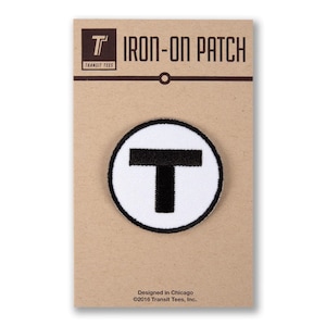 May include: Iron-on patch with a white circle and a black "T" inside. The patch is on a brown card with the text "IRON-ON PATCH" and "TRANSIT TEES" in black. The text "Designed in Chicago ©2016 Transit Tees, Inc." is also on the card.