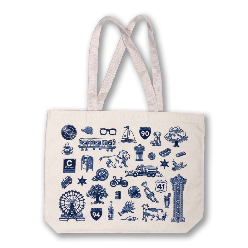 May include: A white canvas tote bag with blue illustrations of Chicago landmarks, including the Chicago "L" train, a hot dog, a Ferris wheel, and the Wrigley Field marquee. The bag has two long handles.